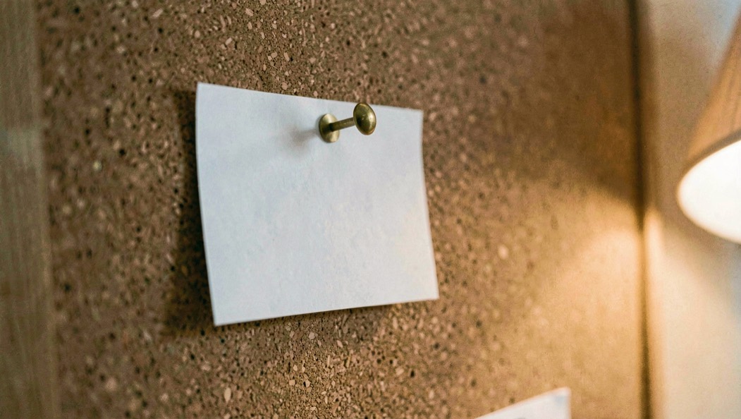 A blank white index card pinned by one brass pin to an otherwise empty corkboard, shallow angle, warm lamp light from the right