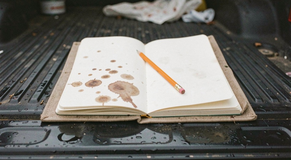 An open cloth bound notebook on a truck tailgate, a pencil laid across the page, dried rain spots on the cover, flat overcast light