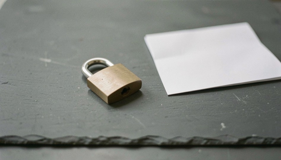 A single closed padlock resting on a plain grey slate surface next to a folded paper sheet, even diffuse light, nothing else in frame