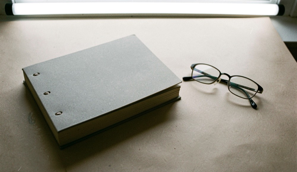 A short stack of plain grey stapled booklets with blank covers on a bare table beside a pair of folded reading glasses, flat overhead light